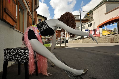 Store front of a hair dressing shop shows a mannequin in a radical pose intended to draw lady patrons to their store who will want a makeover and pay for the service (How Do I Look?).…